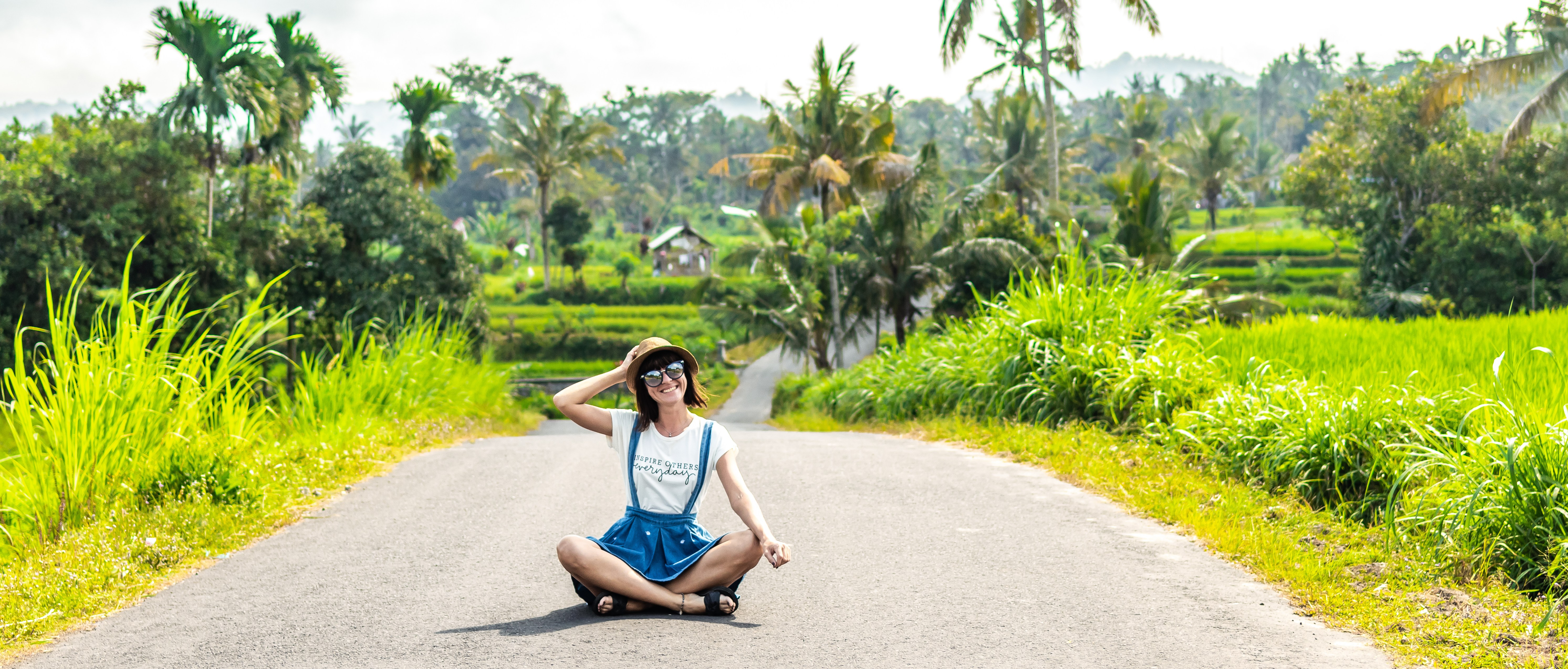 Tropical portrait of young happy woman with straw hat on a road with coconut palms and tropical trees. Bali island.