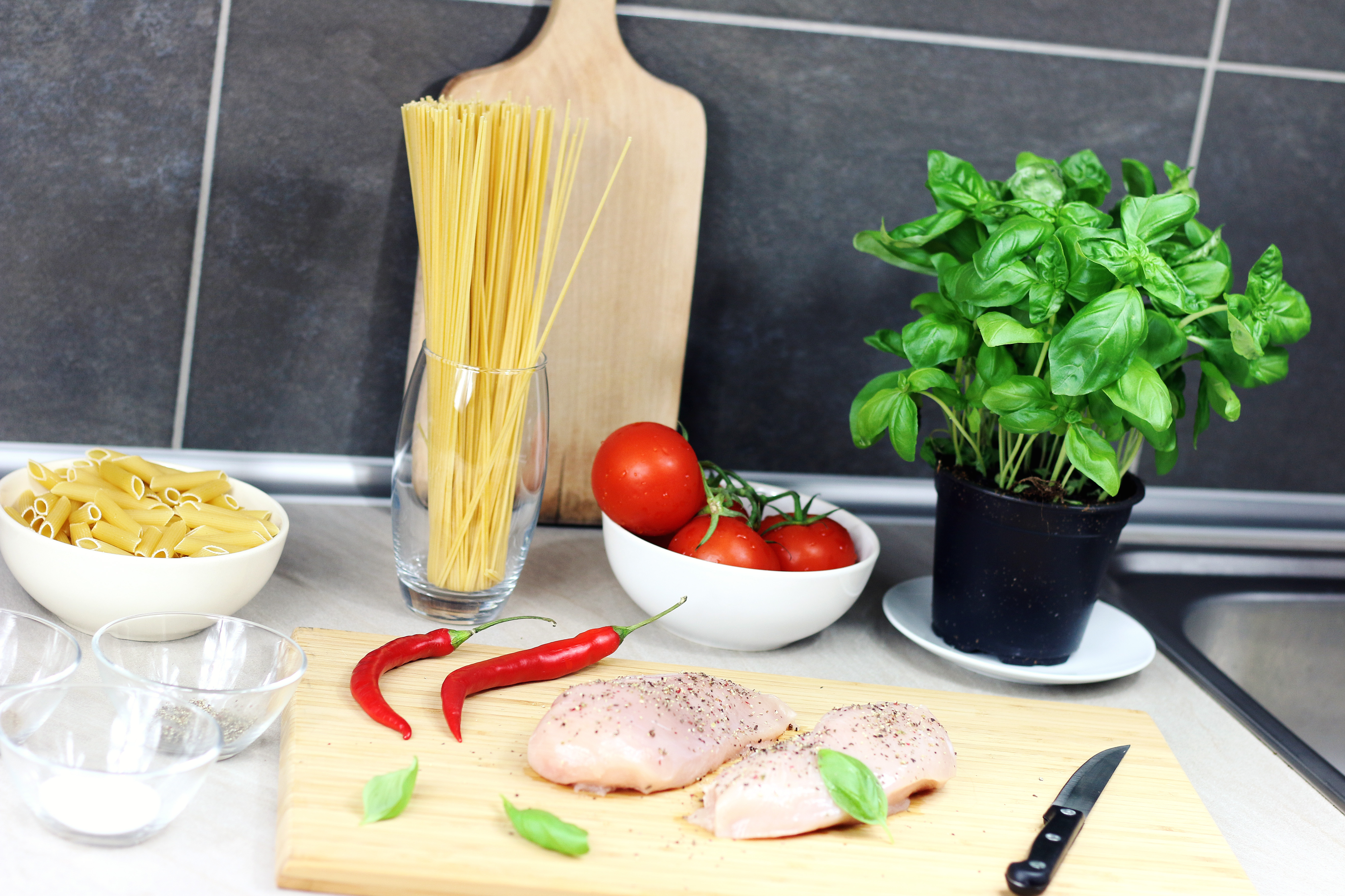 Canva - Raw Chicken Breast Seasoned With Peppers Beside Red Chili, Basil, Bowl of Tomatoes, and Raw Pastas on Table